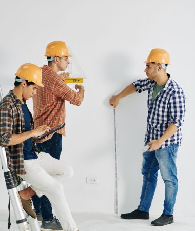 Construction workers measuring wall with various tool when their colleague enterring data in document on tablet computer